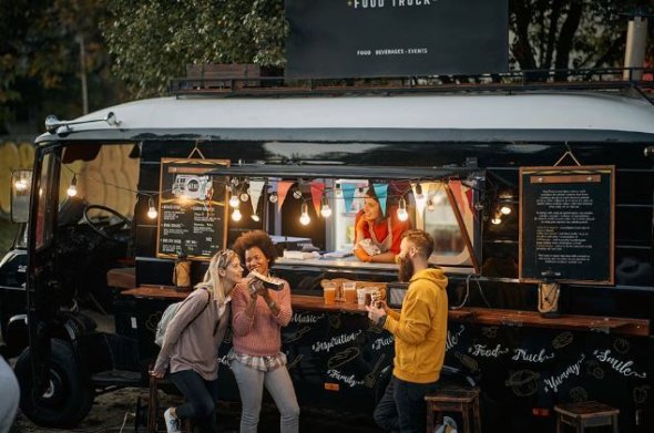 Handel obwoźny w formie food trucka z podświetlaną witryną sprzedażową, trzy osoby zamawiające i rozmawiające na zewnątrz.