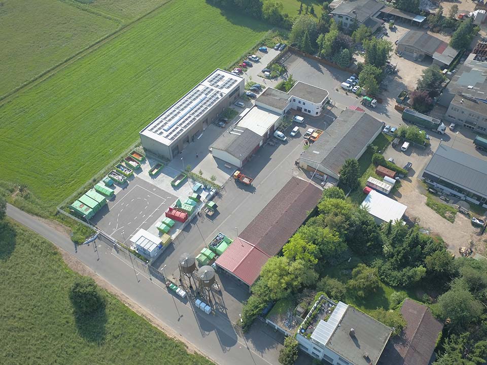 Centro de reciclagem de serviços públicos Vista aérea de um centro de reciclagem com vários edifícios, lugares de estacionamento e contentores de lixo de cores diferentes. Rodeado por prados e árvores.