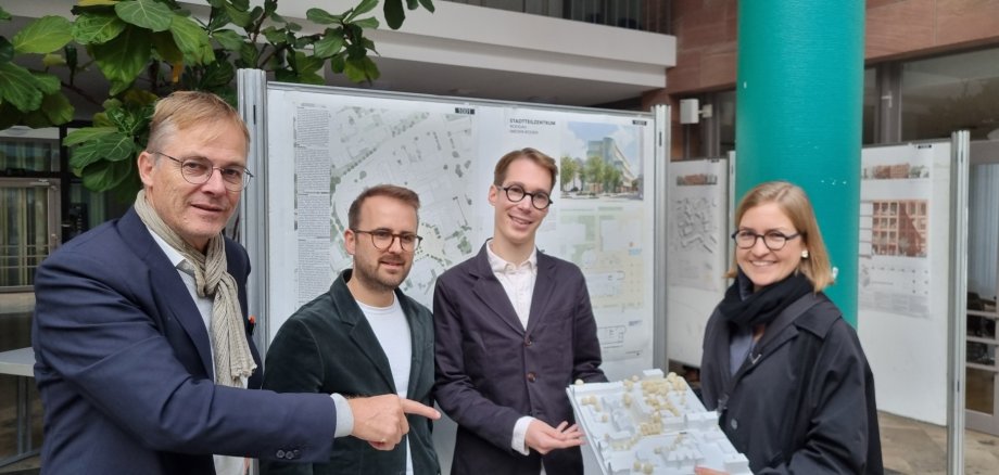 Quatro pessoas, da esquerda para a direita, Marcus Hille (presidente do júri), Valentin Martin, Fabian Wieczorek e Pauline Rohländer (do ROHWAREIMSTUDIO), estão em frente aos planos arquitectónicos do projeto com o modelo vencedor.