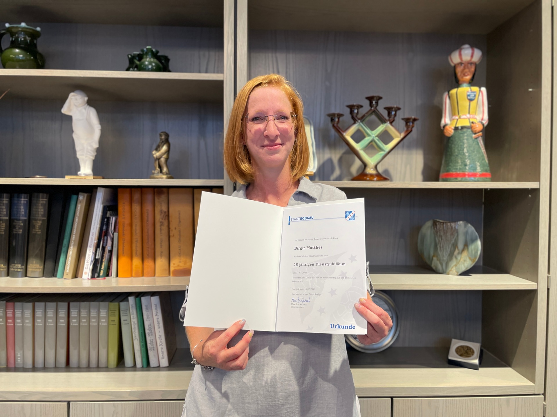 Birgit Mattes celebra 25 anos de serviço Birgit Mattes segura um certificado com a inscrição "25 anos de serviço". Em segundo plano, encontram-se livros, uma estátua e uma vitrina decorativa.