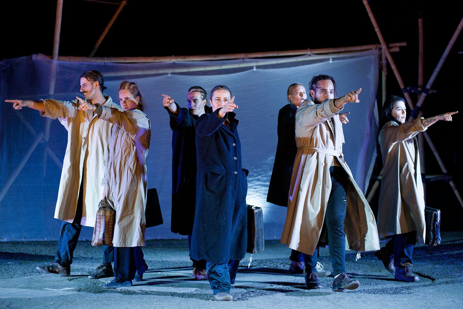 Teatro de rua debaixo das árvores O palco é azul escuro. Seis pessoas com casacos compridos e de cor clara estão em fila e apontam os dedos em diferentes direcções. Algumas seguram sacos nas mãos.