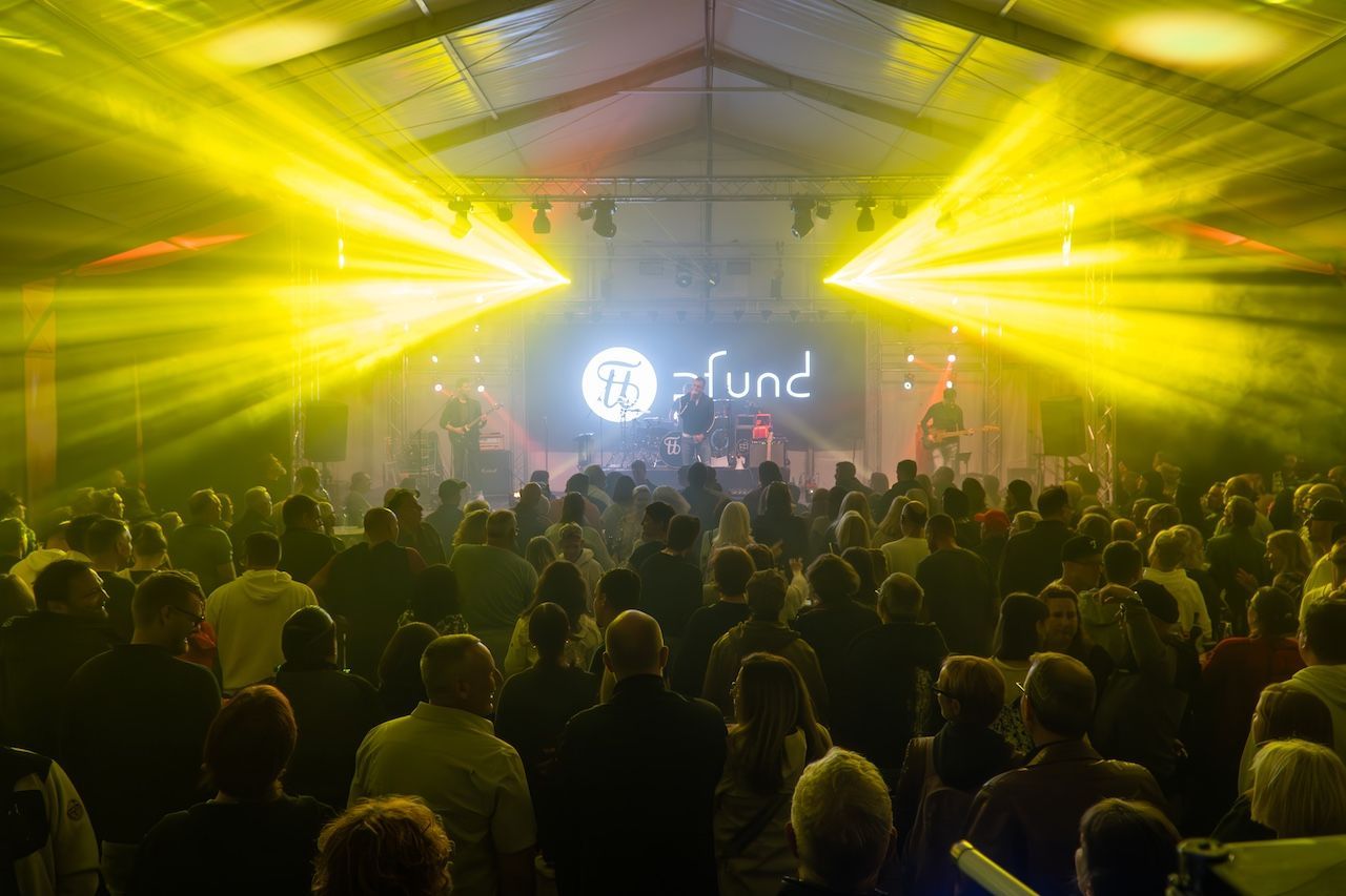 Festival da Indústria Aberta de Rodgau Uma multidão está numa tenda e assiste à atuação de uma banda num palco. Feixes de luz amarela brilham do teto para o público.