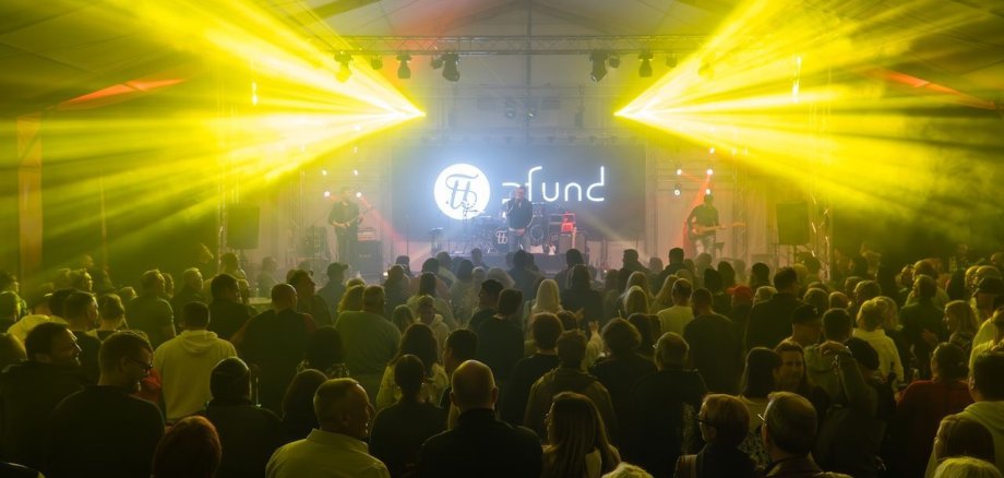 Festival da Indústria Aberta de Rodgau Uma multidão está numa tenda e assiste à atuação de uma banda num palco. Feixes de luz amarela brilham do teto para o público.