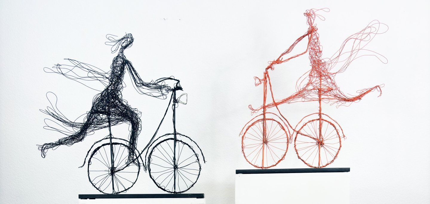 Obra de arte de Henriette Tomasi Duas esculturas delicadas em arame, representando figuras em bicicletas, sobre pedestais brancos, em frente a uma parede branca