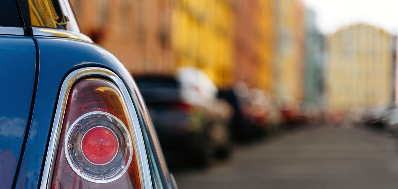 Estacionamento em pavimento Em primeiro plano, vê-se o farolim traseiro de um carro e, ao fundo, um desfiladeiro com casas em frente das quais estão estacionados muitos carros.