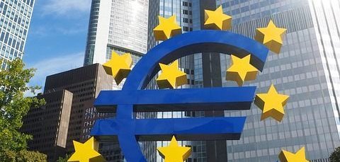 visita guiada vhs Bundesbank Uma grande escultura azul do Euro com estrelas amarelas está em primeiro plano. Ao fundo, vêem-se arranha-céus modernos e um céu azul.