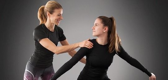 vhs Rückenfit Duas mulheres com roupa de desporto. A mulher da esquerda veste uma T-shirt preta e umas leggings com um padrão roxo. A mulher da direita veste um top preto e umas leggings pretas, com sapatos cor-de-rosa. A mulher da esquerda está a apoiar a mulher da direita, que está numa pose desportiva.