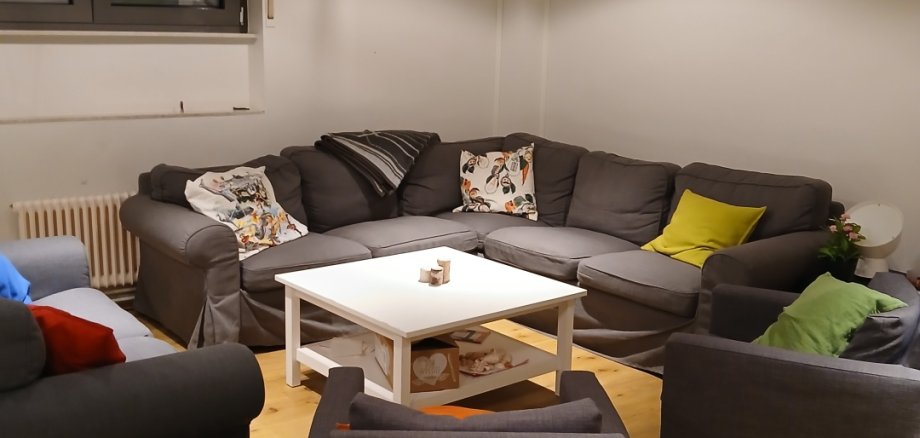 Nesta sala, há um sofá de canto cinzento e três poltronas. Têm muitas almofadas coloridas e uma mesa branca.