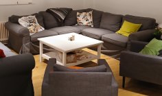 Nesta sala, há um sofá de canto cinzento e três poltronas. Têm muitas almofadas coloridas e uma mesa branca.