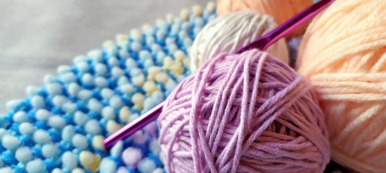 Novelo de lã com agulha de tricotar Três novelos de lã de cores diferentes (roxo, branco, laranja) sobre um fundo azul texturado com uma agulha de croché roxa.