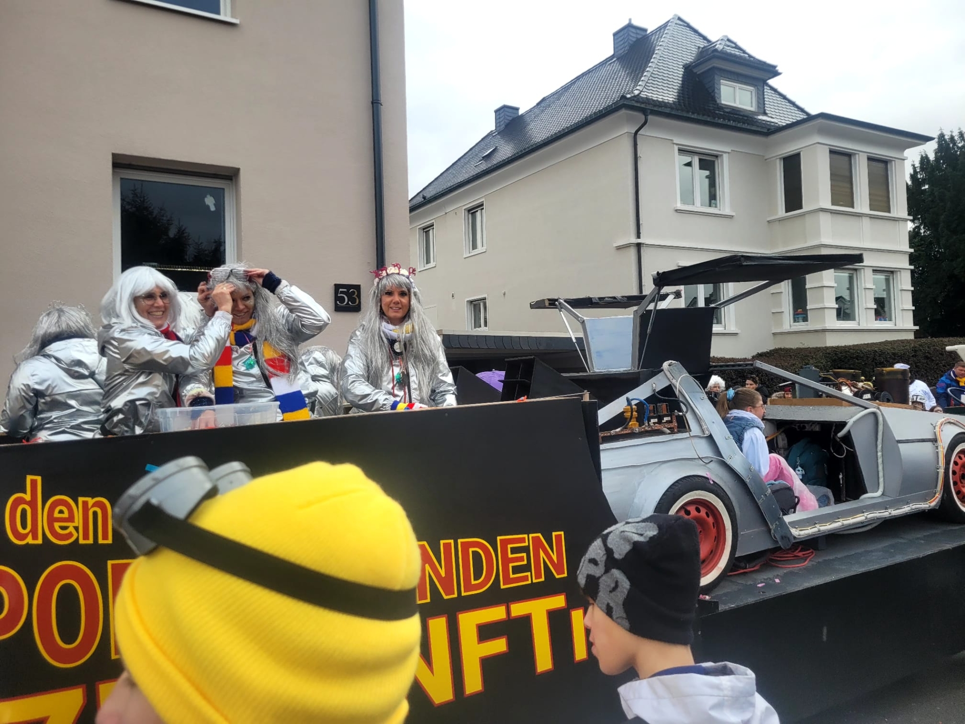 Desfile de Carnaval de 2026 com um carro alegórico que mostra uma réplica do DeLorean do filme "Regresso ao Futuro", pessoas com fatos espaciais prateados e uma criança com um boné amarelo e preto em primeiro plano.