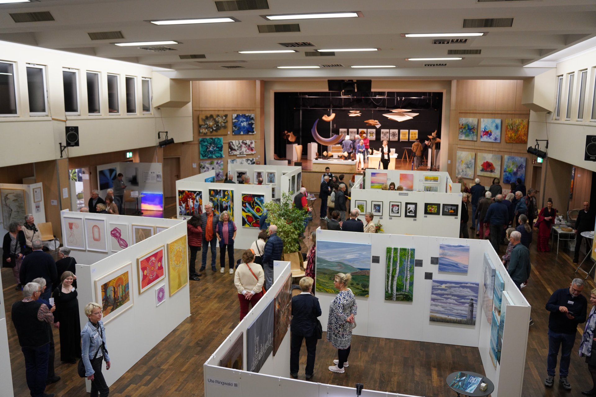 RodgauArt Os visitantes vêem pinturas e esculturas numa sala de exposições na RodgauArt, com obras de arte em paredes brancas e um palco ao fundo