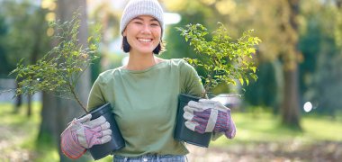 Jardinagem Uma mulher com roupa de jardinagem segura plantas em vasos. Está de pé num parque com árvores ao fundo