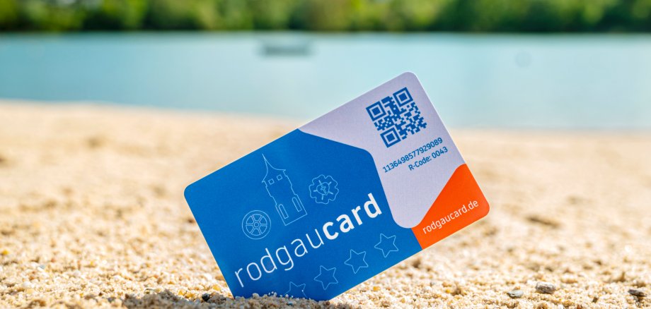 rodgaucard na praia Cartão Rodgaucard espetado na diagonal na areia do lido, com água desfocada e vegetação costeira verde ao fundo