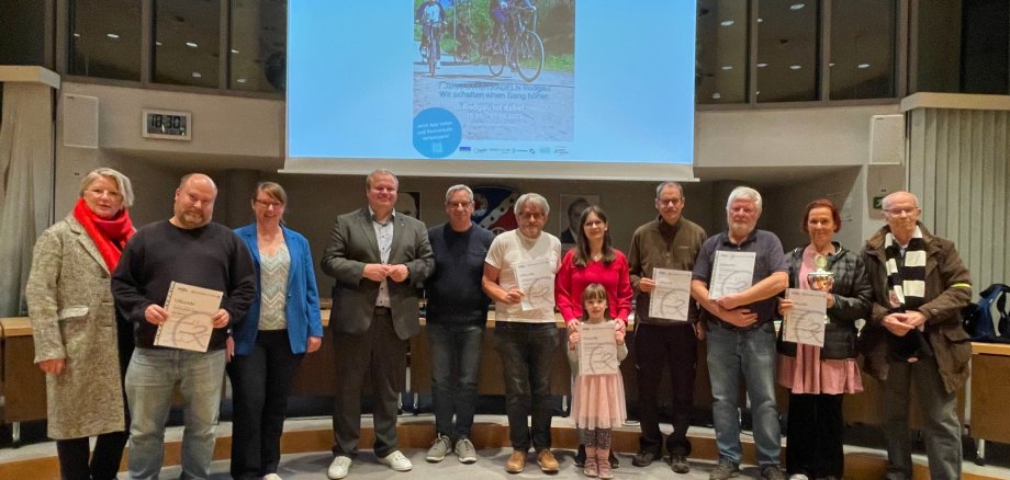 Grup de douăsprezece persoane în picioare pe o scenă, unele ținând certificate, în fundal o imagine de proiector cu motive de biciclete și text despre evenimentul de închidere CITY CYCLING.