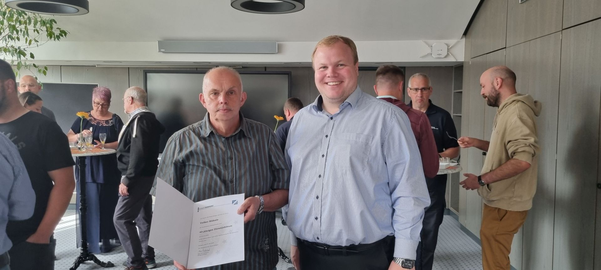 În prim-plan se află niște bărbați, unul dintre ei ținând un document, iar în fundal mai multe persoane care discută într-o atmosferă relaxată, într-o încăpere modernă cu pereți gri.
