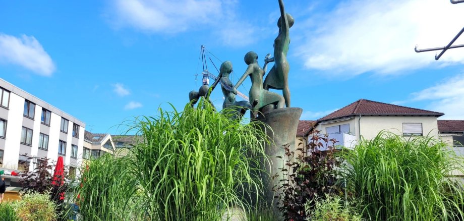 Fântână cu vegetație din Piața Puiseaux Fântână în Piața Puiseaux, cu o sculptură compusă din mai multe figuri, înconjurată de iarbă înaltă și plante, cu clădiri rezidențiale și comerciale în fundal