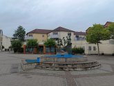 Fântână cu o sculptură cu mai multe figuri pe Puiseauxplatz, înconjurată de copaci și clădiri rezidențiale cu mai multe etaje