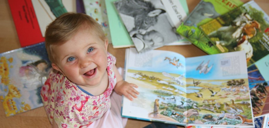 Un copil mic stă pe podea și se uită la mai multe cărți pentru copii deschise, cu ilustrații colorate