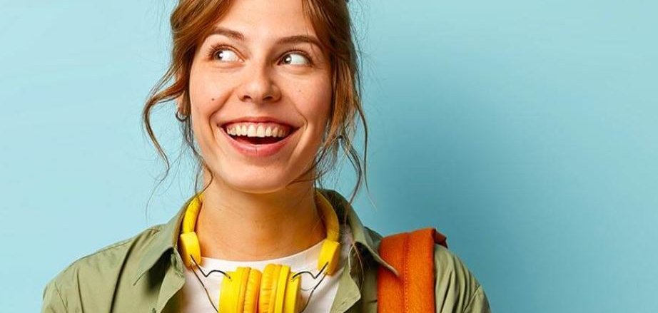 Tânăr cu căști galbene în jurul gâtului, jachetă verde și rucsac portocaliu îndreptat spre stânga