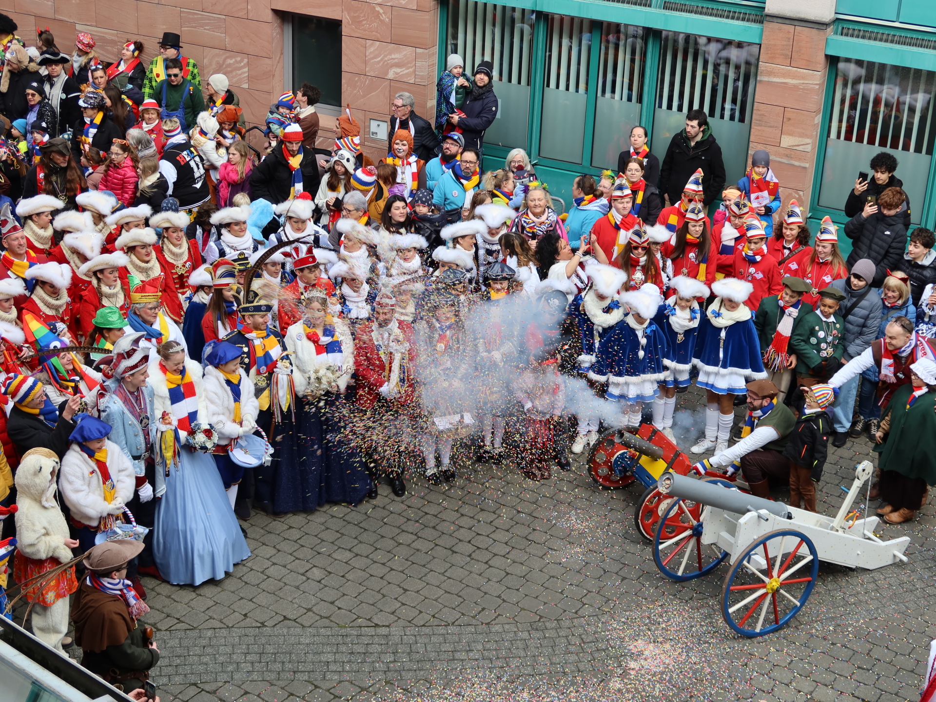 Scenă de grup în timpul carnavalului din fața primăriei. Oameni în costume colorate, unii cu pălării și pene, sărbătoresc și aruncă confetti. Un bărbat îndreaptă un tun de confetti în direcția ușii primăriei.
