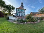Casa Rectorului Geissler cu grădină Rektor-Geißler-Haus din Dudenhofen, o casă istorică cu o jumătate de lemn, cu obloane verzi și albe și un turn, înconjurată de o grădină cu flori și o bancă albă.