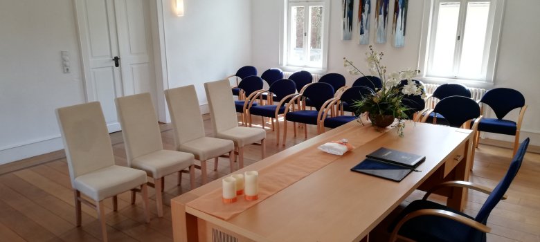 Sală de nunți în Rektor-Geissler-Haus Sală de nunți în Rektor-Geißler-Haus din Dudenhofen cu o masă din lemn deschis la culoare, patru scaune bej, mai multe scaune albastre pe pereți, două ferestre și artă modernă pe perete.