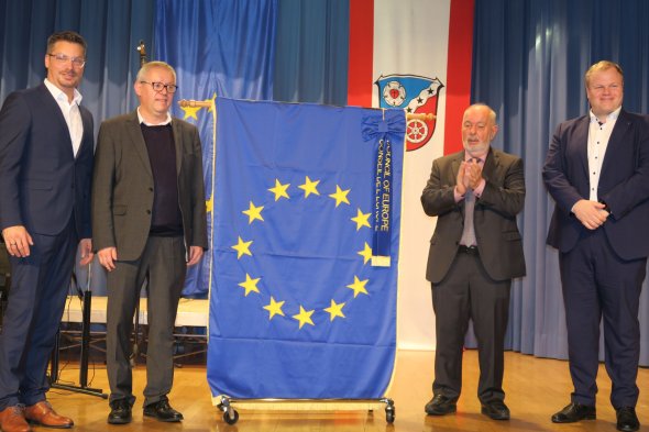 Patru bărbați în costume stau pe o scenă lângă un steag european albastru cu douăsprezece stele galbene. Primarul se află în dreapta. În fundal se află un steag cu o stemă.