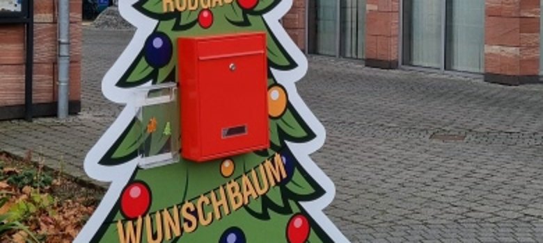 Placa colorată în formă de pom de Crăciun cu o cutie poștală roșie, inscripționată "RODGAUER WUNSCHBAUM", se află pe o piață pavată în fața clădirii primăriei.