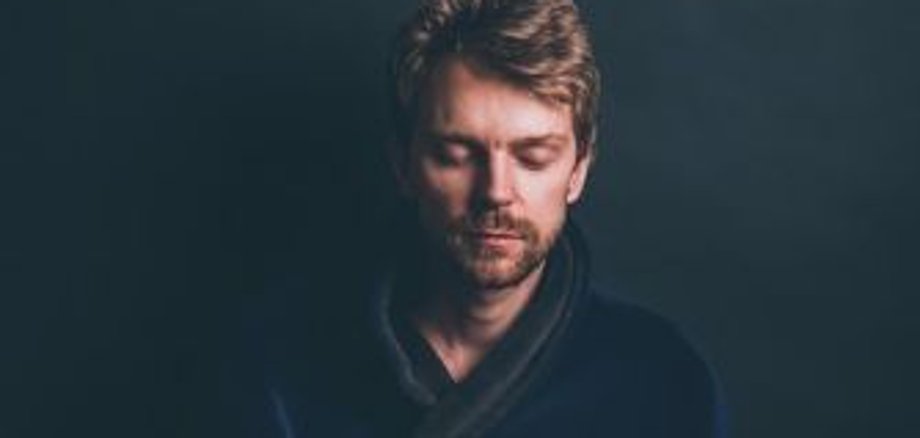 Сердечная чакра Человек сидит, скрестив ноги, на подушке, руки перед сердечной чакрой в позе мудры, темная одежда, темный фон.