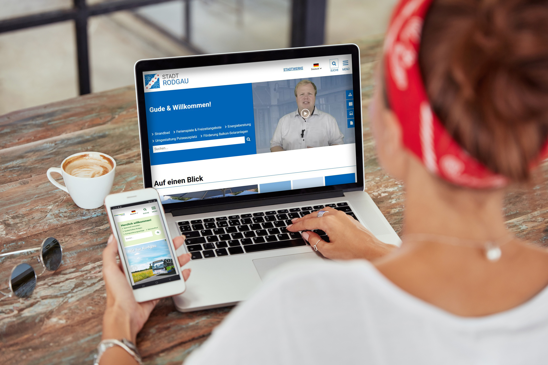 Rodgau şehrinin yeni web sitesi Bir kişi ahşap bir masada oturmuş dizüstü bilgisayar ve akıllı telefon kullanıyor. Dizüstü bilgisayar ekranında Rodgau kasabasının web sitesi ve "Gude & Willkommen!" yazısı görülmektedir. Dizüstü bilgisayarın yanında bir fincan kahve var.