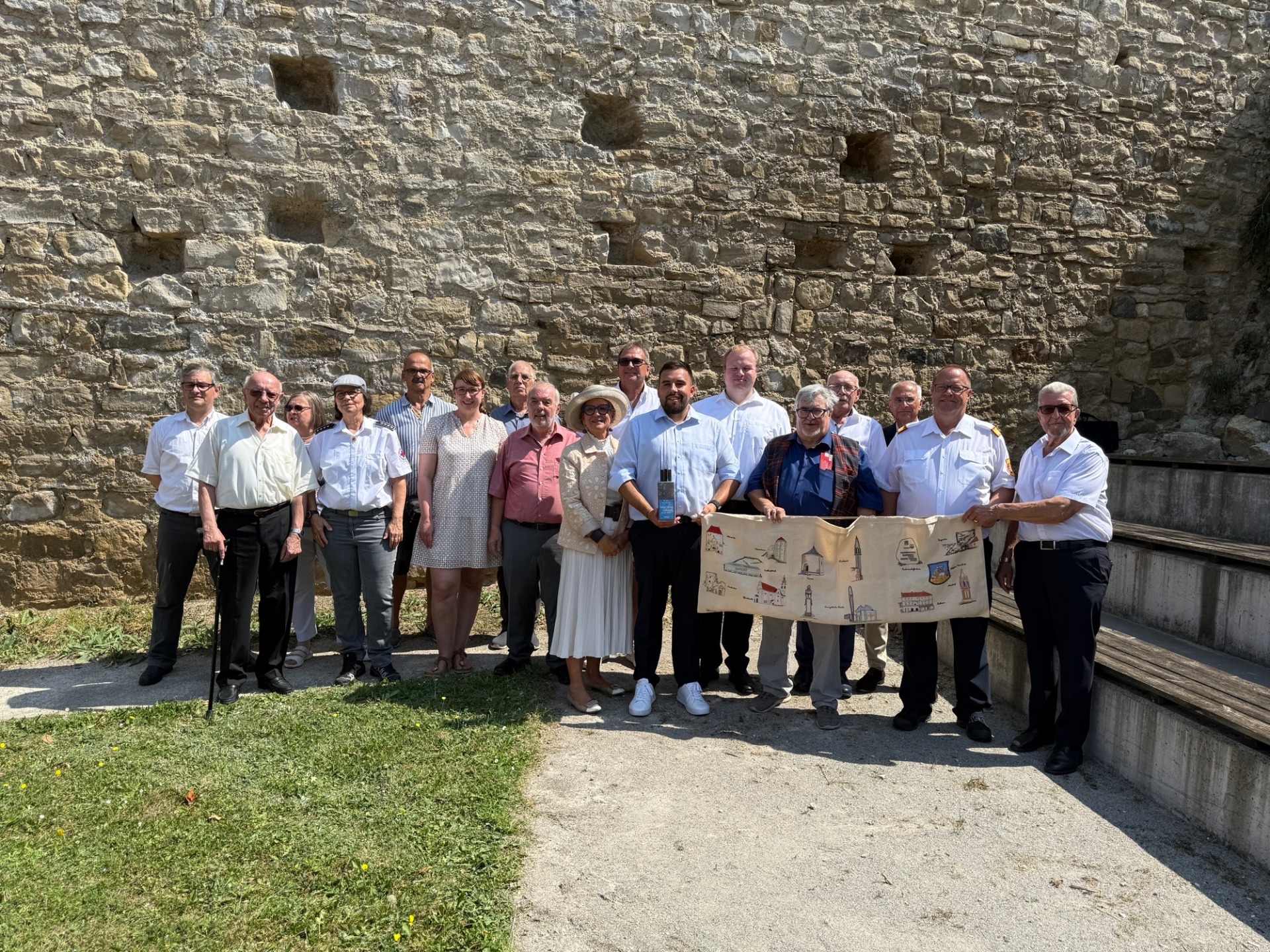 Ortaklık ziyareti 14 kişilik bir grup taş bir duvarın önünde duruyor. Bazılarının elinde üzerinde çizimler olan büyük, katlanmış bir kağıt parçası var. Kıyafetler resmi ve gündelik arasında değişiyor.