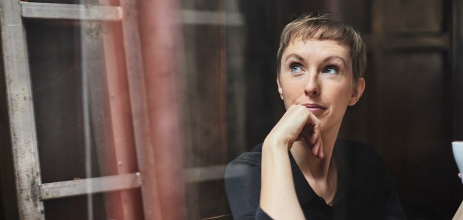 Kısa, sarı saçlı bir kişi karanlık bir odada oturmaktadır. Siyah bir bluz giymekte ve sağ elinde bir fincan tutmaktadır. Arka planda ahşap bir merdiven ve bir duvar görülüyor.