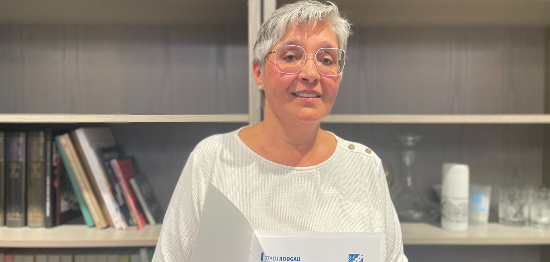 Hona Szycharz Yıldönümü Bir kişi elinde 'Hona Szycharz 40. hizmet yıldönümü' yazılı bir sertifika tutuyor. Arka planda kitap rafları ve bir vazo görülüyor.