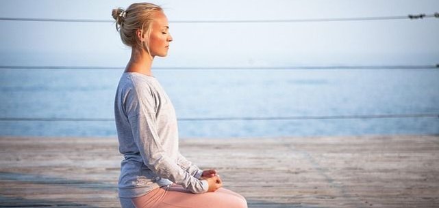 Bir kişi su kenarındaki ahşap bir güvertede lotus pozisyonunda oturuyor. Gri bir üst ve pembe bir tayt giyiyor. Arka planda sakin bir su ve berrak bir gökyüzü görülüyor.