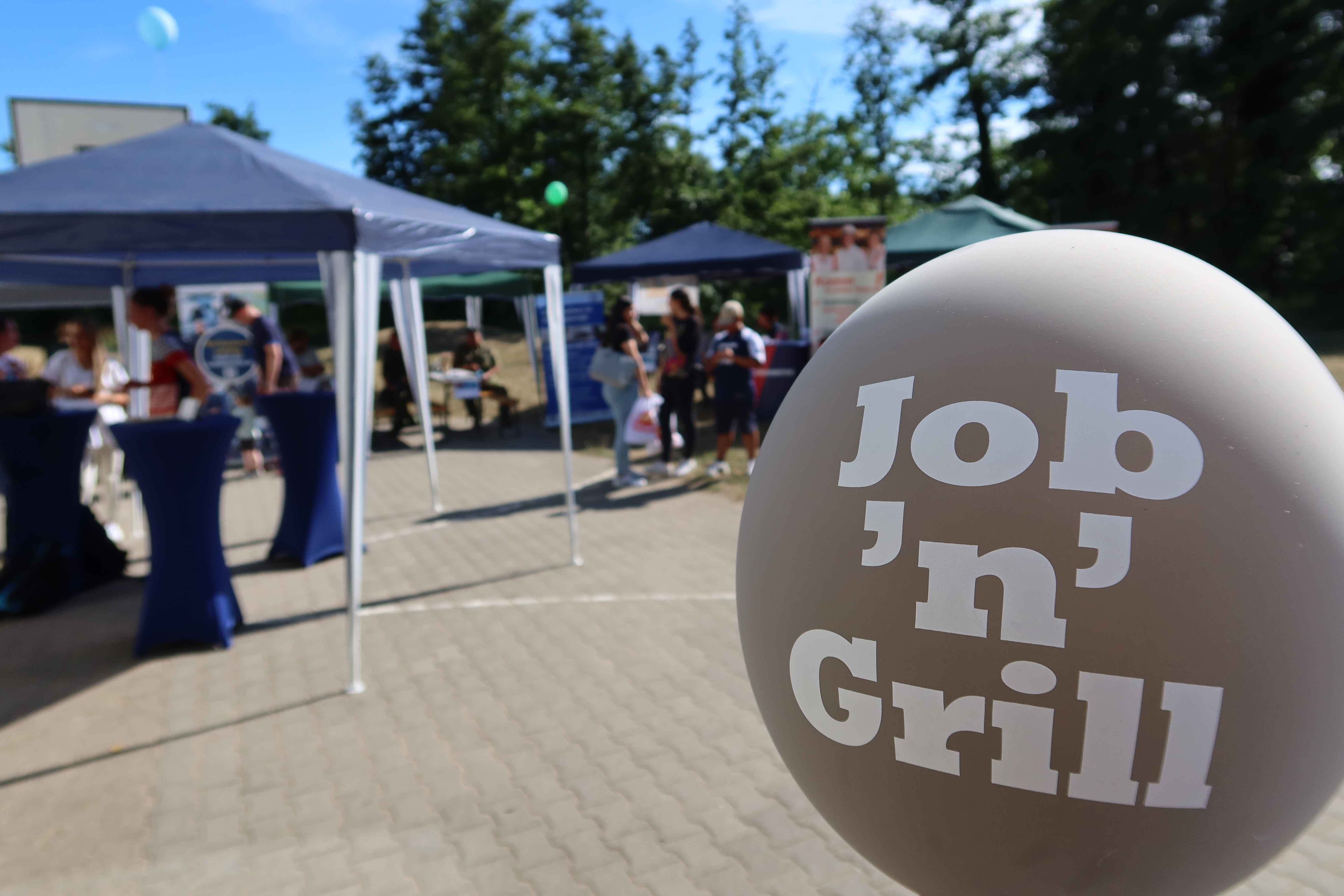 Bir açık hava eğitim fuarında stantların ve ziyaretçilerin önünde 'Job 'n' Grill' etiketli balon