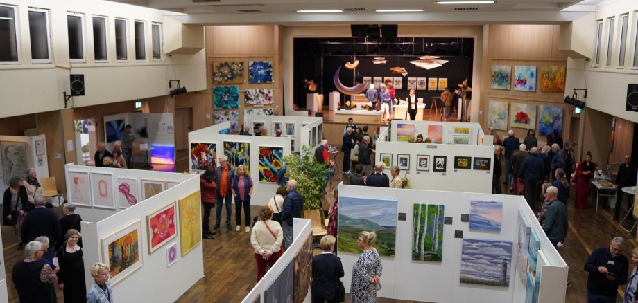 Ziyaretçiler RodgauArt'ın sergi salonunda resim ve heykelleri inceliyor; beyaz duvarlarda sanat eserleri ve arka planda bir sahne yer alıyor