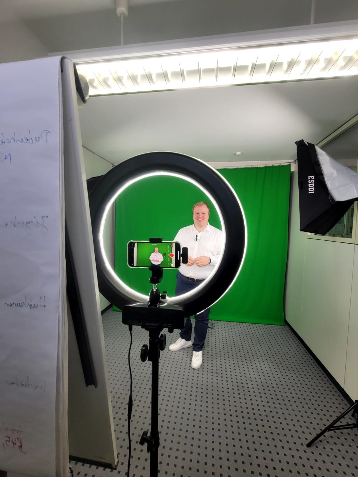 Belediye Başkanı Max Breitenbach beyaz gömleği ve koyu renk pantolonuyla yeşil bir arka planın önünde duruyor ve stüdyo aydınlatmasıyla çevrili bir tripod üzerinde akıllı telefonla halka ışıkla fotoğraflanıyor.