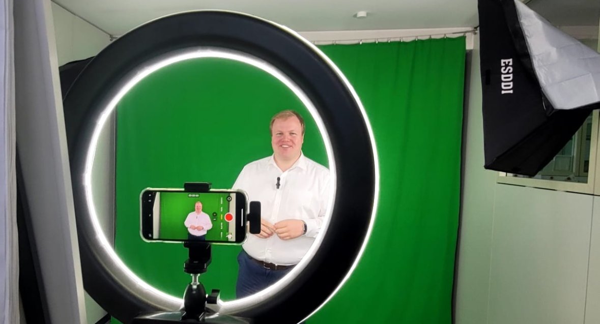 Belediye Başkanı Max Breitenbach beyaz gömleği ve koyu renk pantolonuyla yeşil bir arka planın önünde duruyor ve stüdyo aydınlatmasıyla çevrili bir tripod üzerinde akıllı telefonla halka ışıkla fotoğraflanıyor.