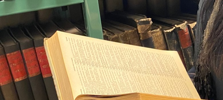 Arşiv kullanımı Kişi kütüphanede eski arşiv ciltlerinin bulunduğu rafın önünde açık bir kitap tutuyor ve okuyor.