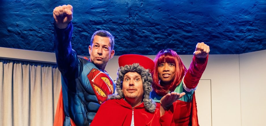 Троє людей на сцені: один у червоному плащі та сивій перуці, двоє в костюмах супергероїв з червоними плащами, кожен з яких тримає перед собою кулак.