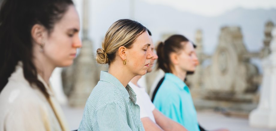 Чотири людини сидять в ряд один біля одного, всі в профіль, перед кам'яною стіною з декоративними елементами, зовні, і медитують.
