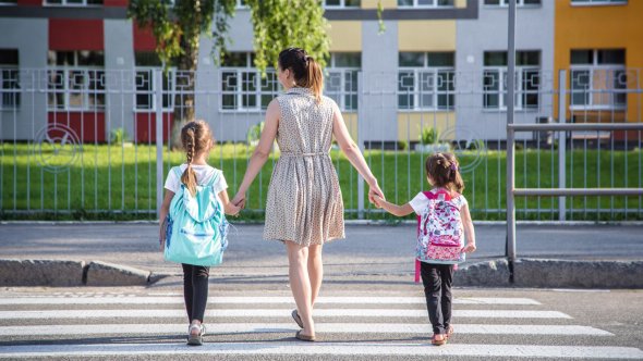 Жінка тримає за руки двох дівчаток з рюкзаками під час переходу по зебрі перед барвистим житловим будинком