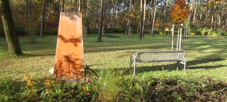 Der Blick richtet sich auf eine Rasenfläche. Links ist eine Stele aus gerostetem Stahl, rechts ein Bank. Im Hintergrund sieht man Kiefern. 