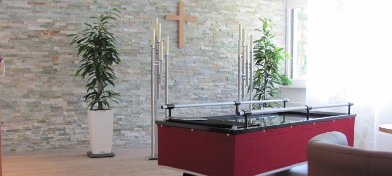 Verabschiedungsraum mit Wagen Im Hintergrund ist eine Steinwand mit Kreuz, davor steht ein Wagen, auf dem ein Sarg aufgebahrt stehen kann.