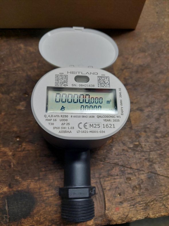 Ein Funkwasserzähler liegt auf hölzernem Untergrund mit aufgeklapptem Display, auf dem eine Reihe von Zahlen und technische Angaben zu sehen sind.