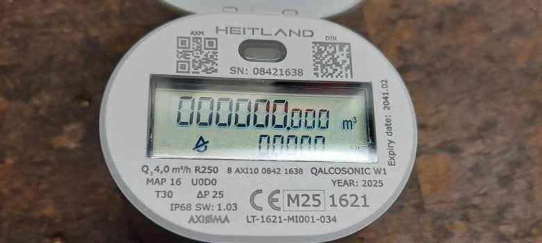 Funkwasserzähler Ein Funkwasserzähler liegt auf hölzernem Untergrund mit aufgeklapptem Display, auf dem eine Reihe von Zahlen und technische Angaben zu sehen sind.